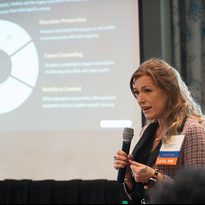 Presenter speaking into a microphone during a conference presentation, with a slide featuring a circular diagram displayed in the background.