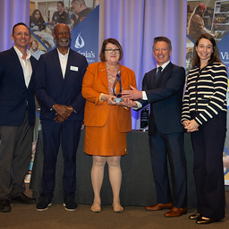 Five award recipients and presenters pose on stage holding a glass award, with Virginia workforce and community college event banners in the background.