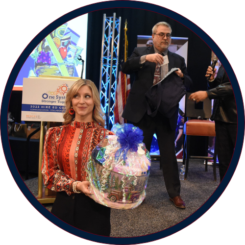 Conference attendee holds a gift basket during a raffle drawing on stage, while event staff select a winner beside presentation screens and event signage.