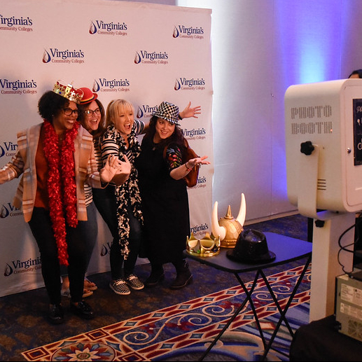 Attendees wearing photo booth props pose in front of a Virginia’s Community Colleges backdrop while a photo booth captures their picture at an indoor event.